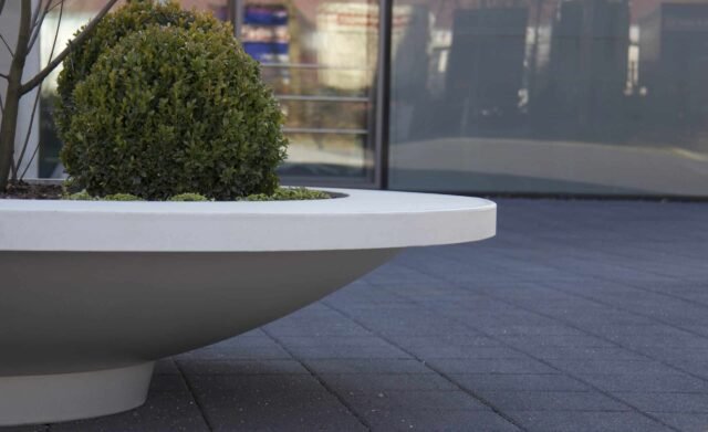 Eine saubere, urbane Szene zeigt die Pflanzschale aus Beton Rund 221cm x H 41cm MATTEO RING in Weiß, mit einem ordentlich geschnittenen Rundstrauch und Grünzeug auf dunkelgrauen Pflastersteinen vor spiegelnden Gebäudefenstern.