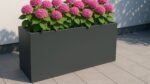 Der Pflanzkübel aus Aluminium Rechteckig 150cm x 50cm x H 60cm EDOARDO 3 in Dunkelgrau steht auf einer gefliesten Terrasse und hält üppig grüne Blätter und blühende rosa Hortensien vor eine helle Wand mit sanftem Sonnenlicht und weichen Schatten.