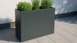 Ein Pflanzkübel aus Aluminium Rechteckig 90cm x 30cm x H 80cm EDOARDO 1 jede RAL Farbe steht auf einer gefliesten Terrasse an einer weißen Wand, die mit Kräutern und grasähnlichen Pflanzen bepflanzt ist. Die mattgraue Oberfläche und das Sonnenlicht schaffen eine ruhige, moderne Atmosphäre im Freien.