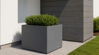 Ein 140x140x80 cm ALESSIO 8 Pflanzkübel aus Aluminium in mattgrau steht auf hellgrauen Fliesen neben einem modernen Gebäude; darin wächst ein dichter grüner Strauch, umgeben von Gras und Kies im Hintergrund.