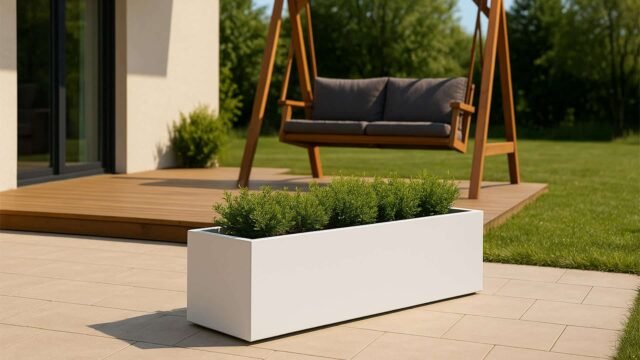 Auf einer modernen Terrasse mit hellen Fliesen steht der Pflanzkübel aus Aluminium Rechteckig 200cm x 50cm x H 60cm EDOARDO 4 jede RAL Farbe, gefüllt mit grünen Sträuchern. Im Hintergrund sind eine hölzerne Veranda, eine zweisitzige Schaukel mit grauen Kissen und ein grasbewachsener Hof mit Bäumen zu sehen.