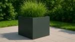 Ein großer, dunkelgrauer Pflanzkübel aus Aluminium Quadratisch 120cm x 120cm x H 80cm ALESSIO 7 steht auf hellgrauen Fliesen, bepflanzt mit hohem, dichtem grünen Gras. Im Hintergrund sind üppige grüne Büsche und Rasen bei Sonnenschein zu sehen.