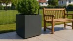 Ein grüner Strauch im Pflanzkübel aus Aluminium Quadratisch 80cm x 80cm x H 80cm ALESSIO 4 (jede RAL Farbe) sitzt auf einer gefliesten Terrasse neben einer Holzbank, mit Drahtzaun, Rasen und fernen Häusern unter einem klaren Himmel.