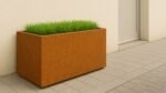 Ein rostfarbener Pflanzkübel aus Cortenstahl Rechteckig 120cm x 50cm x H 60cm CUBI 11 mit hohem grünen Gras steht auf einer hellgrau gefliesten Terrasse neben einer cremefarbenen Wand und einer geschlossenen Tür und verleiht dem modernen, minimalistischen Außenbereich Struktur.