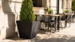Schwarzer trapezförmiger Aluminium-Pflanzkübel mit dichter Thuja neben Tischen auf einer sonnigen Terrasse