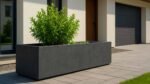 Ein rechteckiger Pflanzkübel aus Beton 40cm x H 40cm SERIE 4 in Grau steht auf einer gepflasterten Terrasse vor einem modernen Haus mit beigen Wänden, Holztür und geschlossener dunkler Garage. In dem Pflanzkübel wächst ein üppiger grüner Strauch.