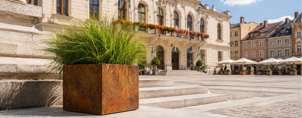Quadratischer Pflanzkübel aus Cortenstahl mit üppigem Ziergras auf einem sonnigen Marktplatz neben dem Rathaus und seitlich an Steinstufen