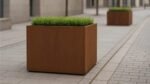 Ein Pflanzkübel aus Cortenstahl Rechteckig 120cm x 80cm x H 100cm CUBI 18 mit Grünpflanzen steht auf einem städtischen Gehweg; ein weiterer identischer Pflanzkübel bereichert den modernen Raum mit seinem geometrischen Pflaster und der urbanen Kulisse.