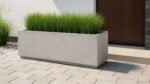 Ein Blumenkübel aus Beton Rechteckig 200cm x 40cm x H 40cm GERALDO 9 in Grau, gefüllt mit hohen grünen Gräsern, steht auf einer gepflasterten Terrasse nahe einem Gebäude mit brauner Tür. Glatte Steine und Ziergräser schmücken die Umgebung.