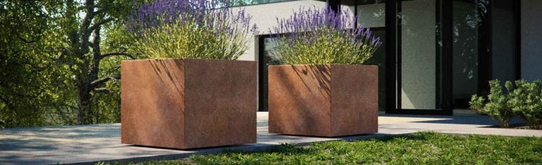 Zwei quadratische Pflanzkübel aus Cortenstahl mit natürlicher Rostpatina, bepflanzt mit Lavendel und Ziergräsern. Die Pflanzkübel stehen auf dezent integrierten Rollen, die von außen kaum sichtbar sind, und befinden sich auf einer modernen Terrasse vor einem zeitgenössischen Gebäude. Die matte, warme Oberfläche des wetterfesten Stahls unterstreicht den modernen Charakter des Außenbereichs.