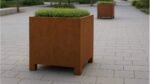 Ein rostfarbener Pflanzkübel aus Cortenstahl Rechteckig 100cm x 80cm x H 100,5cm CECILIA 11 mit grünen Sträuchern steht auf einem hellgrauen Gehweg. Ein weiterer identischer Pflanzkübel steht im Hintergrund, umgeben von üppigem Grün und Bäumen entlang des Gehweges.