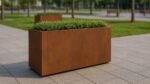 Ein Pflanzkübel aus Cortenstahl Rechteckig 120cm x 50cm x H 70,3cm CECILIA 12 MOBILE mit üppig grünen Sträuchern steht auf einem gefliesten Gehweg. Dahinter bilden Gras, Bäume und eine Holzbank eine städtische Parklandschaft.