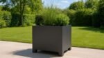 Ein großer, moderner Pflanzkübel aus Aluminium Quadratisch 120cm x 120cm x H 70,5cm APOLLO 8 in einer beliebigen RAL-Farbe steht auf einer hellen Steinterrasse mit hohem Ziergras und einem grünen Strauch; dahinter treffen ein üppiger Rasen und Bäume auf den klaren blauen Himmel.