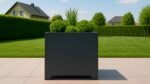Ein großer, moderner Pflanzkübel aus Aluminium mit Füßen Quadratisch 120cm x 120cm x H 110,5cm APOLLO 10 in mattschwarz steht auf einer gefliesten Terrasse mit hohem Gras und runden Sträuchern, vor einem üppigen Rasen, Hecken, Bäumen und einem Haus unter blauem Himmel.