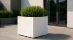 Ein großer, weißer Pflanzkübel aus Aluminium Quadratisch 120cm x 120cm x H 110,3cm APOLLO 10 MOBILE in RAL-Farbe hält einen üppigen Strauch auf einer modern gefliesten Terrasse neben einer Glastür und einer weißen Wand, mit Grün und Gebäudestrukturen im Hintergrund.
