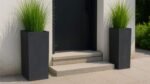 Zwei hohe, schwarze, rechteckige Blumenkübel Beton ALESSANDRO (40x40xH80cm) mit grünem Ziergras flankieren eine kleine, helle Steintreppe zu einer schwarzen Tür an einer weißen Wand und sorgen für einen klaren, modernen Look mit neutralen Fliesen und minimalistischem Design.