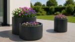 Drei moderne Pflanzkübel aus Aluminium Rund 100 cm x H 47 cm Daria (1 Set, jede RAL-Farbe) sitzen auf einer gefliesten Terrasse, die mit leuchtend rosa, lila und weißen Blüten gefüllt ist. Ein grüner Rasen und Hecken erscheinen im sonnigen Hintergrund.