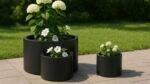 Drei schwarze Pflanzkübel aus Aluminium Rund 80 cm x H 38 cm Daria 2 Set, jeweils mit weißen Blüten und grünen Blättern, sitzen auf einer Steinterrasse vor einer Kulisse aus üppigem Gras und einer ordentlich gestutzten Hecke in einem friedlichen Garten.