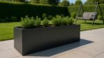 Pflanzkübel Aluminium Groß Rechteckig 200cm x 50cm x H 50cm EDOARDO 7 in jeder RAL Farbe mit grünen Pflanzen steht auf einer gefliesten Terrasse. Dahinter ein Garten mit Rasen, Hecke und grauer Gartenschaukel in sonniger Atmosphäre.