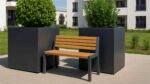 Eine kleine Holzbank mit Metallrahmen sitzt auf dem Bürgersteig zwischen zwei Pflanzkübeln Aluminium Groß Quadratisch 120x120x120cm ALESSIO 15 in RAL-Farbe, gefüllt mit grünen Sträuchern. Im Hintergrund sind moderne weiße Wohnungen und eine Rasenfläche unter blauem Himmel zu sehen.