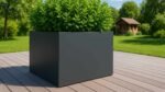 Ein quadratischer, dunkelgrauer Pflanzkübel Aluminium Groß 180cm x 180cm x H 100cm Alessio 17 in einer beliebigen RAL-Farbe mit üppigen grünen Pflanzen steht auf einer Holzterrasse, die von einem grasbewachsenen Rasen, Bäumen und einem kleinen braunen Holzhaus unter einem klaren Himmel umgeben ist.