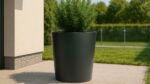 Ein Pflanzkübel Aluminium Groß 100cm x H 100cm Silvestro 3 in mattem Schwarz steht auf einer gefliesten Terrasse an der Hauswand und hält einen üppig grünen Strauch. Dahinter vervollständigen ein sonnenbeschienener Rasen, eine Hecke und ein Zaun die gepflegte Gartenszene.