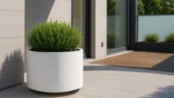 Ein ovaler, weißer Aluminium-Pflanzkübel TIZIANO 1 (∅60cm x H 60cm, jede RAL-Farbe) steht auf einer gefliesten Terrasse neben einer modernen Glastür; Sonnenlicht und Grünpflanzen unterstreichen die moderne, ruhige Atmosphäre im Freien.