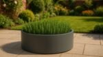 Ein runder, grauer Pflanzkübel aus Aluminium, gefüllt mit aufrechten, grünen Rosmarinpflanzen, steht auf einer gefliesten Terrasse. Im Hintergrund erinnern ein üppiger Rasen und verschwommene Gartensträucher an einen gepflegten Hinterhof oder Garten an einem sonnigen Tag.