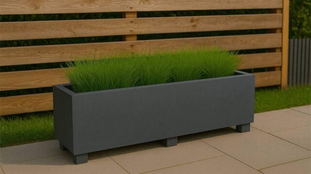 Ein schwarzer Beton Pflanzkübel Rechteckig 200cm x 50cm x H 50cm FRANCESCO, gefüllt mit hohem grünen Gras, steht auf einer gefliesten Terrasse. Dahinter befindet sich ein waagerechter Bretterzaun mit Grünzeug im Hintergrund.