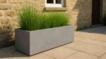 A Beton Pflanzkübel Rechteckig 120cm x 40cm x H 40cm GERALDO in Grau, bepflanzt mit hohen Gräsern, steht auf einer Stein-Terrasse neben einer beigen Wand und unter einem Fenster - für ein modernes, minimalistisches Ambiente.