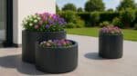 Drei anthrazitfarbene Aluminium-Pflanzkübel in unterschiedlichen Größen mit violetten, rosa und weißen Blüten auf einer sonnigen Terrasse