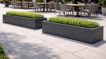 Zwei lange, anthrazitfarbene Beton-Pflanztröge mit schlanker Seitenwand, bepflanzt mit 15 cm hohem grünem Ziergras, platziert neben Terrassen-Tischen bei sonnigem Wetter.