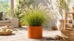 Zylindrisches Pflanzgefäß in Cortenstahl-Optik mit üppigem Ziergras auf einer hellen, sonnigen Terrasse mit natürlichen Gartenaccessoires