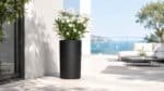 Schwarzer Aluminium-Pflanzkübel Tiziano 6 mit weißen Blüten auf einer hellen Terrasse, 35 cm Durchmesser und 100 cm Höhe