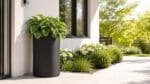 Schwarzer Aluminium-Pflanzkübel Tiziano 9 mit grünen Hostas vor einem hellen Haus, 60 cm Durchmesser und 100 cm Höhe