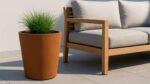 Ein mit hohem grünem Gras bepflanzter Pflanzkübel aus Cortenstahl Rund 50 cm x H 50 cm VASO 10 steht auf einer gefliesten Terrasse neben einem hellen Holzsofa mit grauen Kissen, das in natürliches Sonnenlicht getaucht ist, das knackige Schatten wirft.