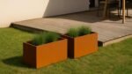 Zwei Pflanzkübel ohne Boden aus Cortenstahl Rechteckig 100cm x 50cm x H 50 cm Corten Frame 11 mit grünem Ziergras stehen auf einem grasbewachsenen Rasen neben einer hellen Holzterrasse, mit einem Stuhl und Tisch in der Nähe und einer weißen Wand im Hintergrund.