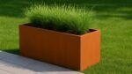 Ein Pflanzkübel ohne Boden aus Cortenstahl Rechteckig 150cm x 50cm x H 60 cm Corten Frame 20 gefüllt mit hohem grünem Ziergras steht auf einem sauber getrimmten Rasen neben hellgrauen quadratischen Fliesen im hellen Sonnenlicht.