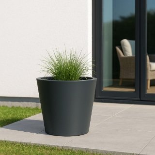Ein großer, dunkelgrauer Aluminium-Pflanzkübel mit hohem grünem Gras steht auf einer gefliesten Terrasse vor einem modernen Haus mit weißen Wänden, einer Glastür und einem Blick auf Korbmöbel. Im Hintergrund sind Gras und ein gepflegter Rasen zu sehen.