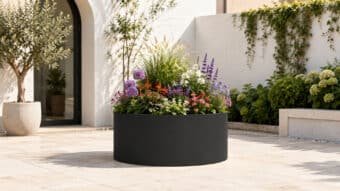 Niedriger, runder Aluminium-Pflanzkübel in Mattschwarz mit farbenfroher Blütenbepflanzung auf einem hellen, mediterranen Innenhof