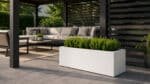 Weiße rechteckige Pflanzkübel mit grünen Sträuchern in einer sonnigen modernen Gartenlaube mit Lounge-Möbeln