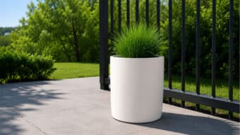 Weiße runde Pflanzkübel mit grünem Ziergras neben einem schwarzen Metallzaun auf einer sonnigen Terrasse