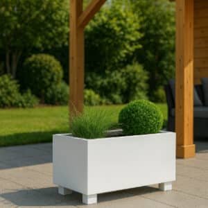 Ein stilvolles Blumenkübel mit Füßen in Weiß mit grünem Gras und rundem Strauch steht auf einer gefliesten Terrasse. Nahebei sind eine Holzpergola und eine Holzkonstruktion, im Hintergrund eine Wiese und Bäume an einem sonnigen Tag.