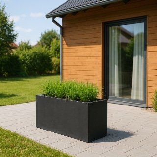 Ein moderner rechteckiger schwarzer Pflanzkübel mit hohem grünen Gras sitzt auf einer grau gefliesten Terrasse vor einem Holzhaus mit großen Glastüren. Die Terrasse ist umgeben von üppigem Gras und Bäumen unter einem klaren, sonnigen Himmel.