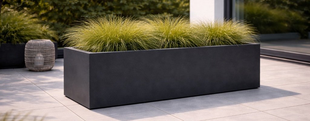 Ein moderner rechteckiger schwarzer Blumenkübel, gefüllt mit hohen grünen Ziergräsern, steht auf grauen Fliesen. Daneben ist eine dekorative graue Laterne platziert, umgeben von üppigem Grün und einem Gebäude mit Glasfenstern im Hintergrund.