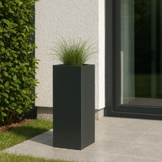 Ein hoher, rechteckiger Pflanzkübel Fiberglas hoch in mattschwarz mit grünem Ziergras steht auf einer grau gefliesten Terrasse neben einer weißen Hauswand und Glastür. Im Hintergrund sind gestutzte Hecken und Rasen an einem hellen, sonnigen Tag zu sehen.