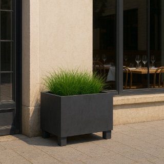 Ein rechteckiger schwarzer Pflanzkübel mit grünem Ziergras dient als stilvoller Sichtschutz auf dem gepflasterten Gehweg vor einer beigen Gebäudewand. Rechts geben große Fenster den Blick ins Restaurant mit gedeckten Tischen und Holzstühlen frei.