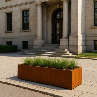 Ein großer rechteckiger Cortenstahl-Pflanzkübel nach Maß mit hohem grünem Gras steht auf einem gepflasterten Weg vor einem stattlichen Steingebäude mit Säulen, Rundbogenportal und Stufen, die zu einer dunklen Holztür führen. Sträucher säumen das Gebäude im Hintergrund.