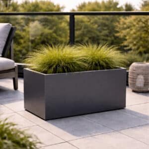 Ein rechteckiger schwarzer Blumenkübel mit dichtem, spitzem Gras steht auf dem gefliesten Patio. Links ein Polsterstuhl, rechts eine Korblaterne und dahinter Bäume - stilvoll inszeniert mit einem Pflanzkübel nach Maß.