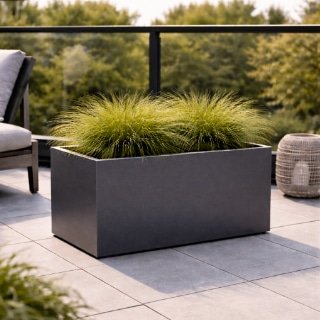 Ein rechteckiger schwarzer Blumenkübel mit dichtem, spitzem Gras steht auf dem gefliesten Patio. Links ein Polsterstuhl, rechts eine Korblaterne und dahinter Bäume - stilvoll inszeniert mit einem Pflanzkübel nach Maß.